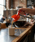 Person pouring coffee from a 64 Oz Commercial Glass Coffee Pot Decanter With Orange Decaf Handle For Restaurants, Cafes, And Offices.
