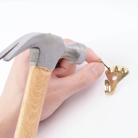 Hand using hammer to attach nail to hanger from DIY Picture Hanging Hardware Kit 220 Piece Brass Finish Set With 24 Ten Pound, 14