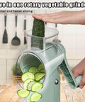Side view of green 5-in-1 rotary vegetable grinder with hand crank; cucumber slices visible.