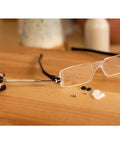 11 Piece Universal Compact Eyeglasses Repair Kit With Screwdriver And Micro Screws Portable Case For Quick Fixes On All Kinds Of Broken Glasses Frames Travel Ready close-up view, including transparent eyeglasses, screwdriver, screws, and nose pads on wooden surface.