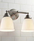 Brushed nickel double wall sconce with frosted bell-shaped glass shades against white subway tile wall.