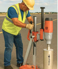 Diamond core drill on a portable orange-gray stand; front view of a worker in safety gear operating the tool.
