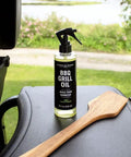 Plant Based BBQ Grill Cleaner Oil For Grates And Grills 8 oz bottle on grill with wooden scraper in outdoor setting