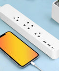 Smart Power Strip With WiFi And Bluetooth Remote Access, Voice Activated Outlets, Quick Charge USB And Type C Ports, App Managed Surge Blocker on a blue surface with a smartphone charging.