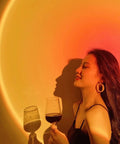 Sunset Lamp Projector With Remote 13 Color Multicolor Night Light casting warm orange light on woman with wine glass.