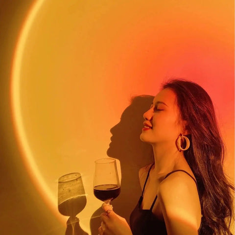 Sunset Lamp Projector With Remote 13 Color Multicolor Night Light casting warm orange light on woman with wine glass.