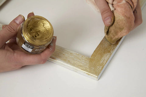 Pebeo Gilding Wax Empire Gold jar held near a gold-painted frame corner being brushed on with cloth