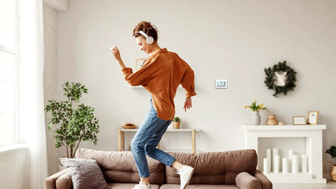 Woman dancing in living room with 7 Day Flexible Programmable Thermostat With Extra Large Backlit Display White visible on wall.