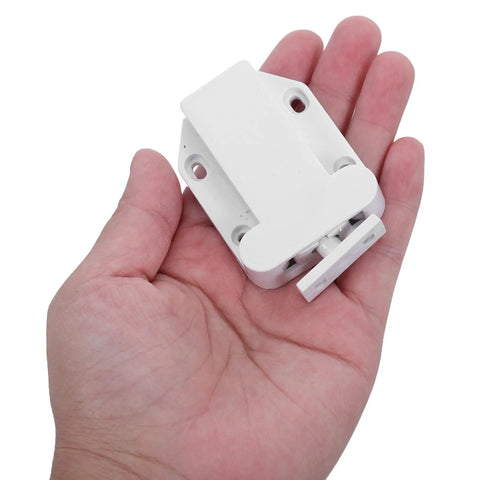 Hand holding one of the 8 Pack No Fuss Self Closing Cabinet Latches With Push Button Door Opener Automatic Rebound Closers in white.