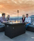44 Inch Outdoor Propane Fire Pit Table With 50,000 BTU Heat, tempered glass top, used by a group sitting on a patio at sunset.