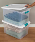 A hand lifting the lid of a clear plastic latching storage bin holding folded blankets, part of the Slay Your Storage Goals With This Badass Set Of Plastic Latching Storage Bins Featuring Secure Snap Lids, Includes 106 Qt And 64 Qt Totes For Closet Decluttering, Garage Organization, And Keeping Bugs At Bay.