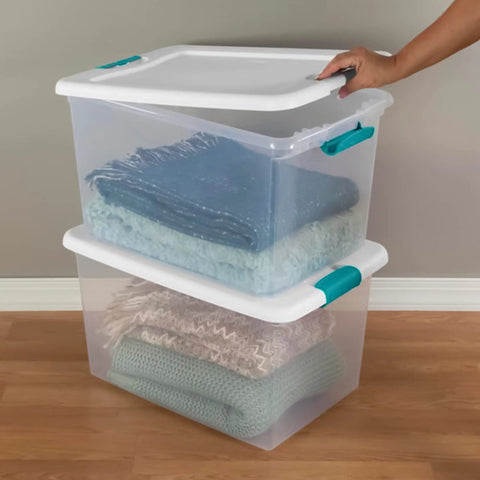 A hand lifting the lid of a clear plastic latching storage bin holding folded blankets, part of the Slay Your Storage Goals With This Badass Set Of Plastic Latching Storage Bins Featuring Secure Snap Lids, Includes 106 Qt And 64 Qt Totes For Closet Decluttering, Garage Organization, And Keeping Bugs At Bay.
