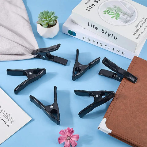 Beefy 7 Pack 4 Inch Spring Clamps Set for arts, crafts, woodworking, backdrops; six black metal clips on a table with plants and books.
