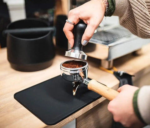 Barista using Coffee Tamper Mat For Portafilter 8x8 Inches Silicone Tamping Pad Non Slip Heat Resistant Barista Accessory For Espresso Machines Easy Wipe Clean Perfect For Home Or Cafe.