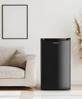 Front view of Upstreman 3.2 cu ft black single-door mini fridge with freezer beside a beige sofa in a living room.