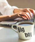 Large white coffee mug with 'Big boy' text beside person typing on laptop in morning light
