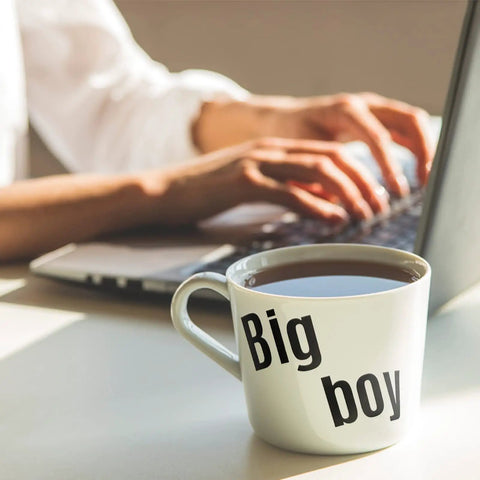 Large white coffee mug with 'Big boy' text beside person typing on laptop in morning light