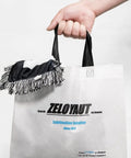 Hand holding a white tote bag with black handles and a logo for ZeloYaut Sublimation Supplier, featuring a fringe decoration on top.