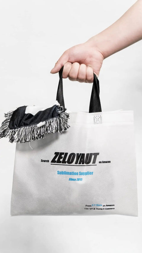Hand holding a white tote bag with black handles and a logo for ZeloYaut Sublimation Supplier, featuring a fringe decoration on top.