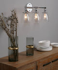 Three Light Industrial Wall Sconce With On Off Switch And Clear Glass Shade For A Cool Retro Vibe Vintage Antique Brass Vanity Lighting For Bathroom And Living Room Dimmable When Paired With A Compatible Dimmer Bell Shade Finish illuminated above wood cabinet