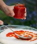 Pizza dough on a glass tray being sauced by a hand holding a jar of tomato sauce.