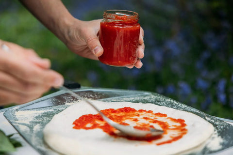Pizza dough on a glass tray being sauced by a hand holding a jar of tomato sauce.