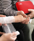 Red I GO mini hard-shell first aid kit with zipper, held beside a child’s bandaged arm.