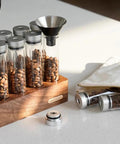 12 Piece Single Dose Coffee Bean Storage Tubes With One Way Exhaust Valve Glass Vials On Sturdy Walnut Wood Base and Easy Pouring Funnel with beans.