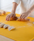 Person rolling dough on a yellow mat with dumplings and eggs nearby