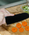 Side view of black pleated crinkle cutter with orange thumb guard, slicing five carrot coins on a wooden cutting board.