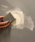 Hand using sandpaper block on wall patch, part of drywall repair kit with tapered hole saw and two plugs for clean flat patches.