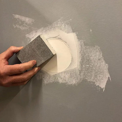 Hand using sandpaper block on wall patch, part of drywall repair kit with tapered hole saw and two plugs for clean flat patches.