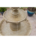 Close-up of water flowing in a beige stone tiered fountain on a patio, surrounded by potted plants and brick flooring.