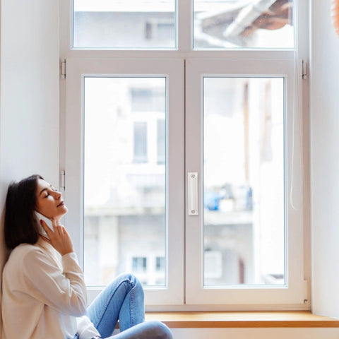 Woman seated by a modern window, enjoying a phone call in a well-lit room with a serene view outside.