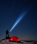 Silhouette holding a Two Cell D LED Flashlight With Adjustable Beam QuickFocus System near a glowing tent under a starry sky.