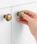 Round Antique Brass Cabinet Knobs 1-1/4 Inch, 10 Pack, Beefy Heavy Duty Metal Hardware With Rust Resistant Finish, Includes Two Screw Sizes 1 Inch And 1-1/2 Inch For Easy Installation On Kitchen And Bathroom Cabinets. Hand holding round antique brass cabinet knob on white cabinet door.