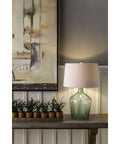 Hatteras Hammered Glass Table Lamp In Seabreeze Blue With White Shade on a wood console, next to potted plants and artwork