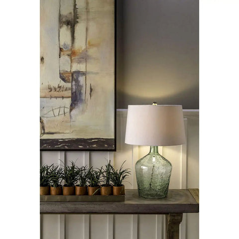 Hatteras Hammered Glass Table Lamp In Seabreeze Blue With White Shade on a wood console, next to potted plants and artwork
