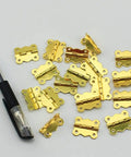 20 Pack Small Cabinet Door Hinges For Furniture Upgrade Heavy Duty Drawer Mounting Braces Smooth Swing Closet Cupboard Fittings Built Like A Tank Ultra Durable Steel Perfect For Diy Renovations And Home Makeovers. Gold small cabinet door hinges on white background with pen.