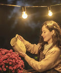 Woman watering flowers outdoors under string lights with Outdoor Smart Plug With Two Outlets WiFi Outlet That Works With Alexa Google Home And IFTTT No Hub Required IP64 Weather Resistant Sunset Sunrise Offset Control Two Outdoor Devices From Anywhere Up To 300 Feet.