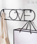 Sleek Wall Mounted Coat Rack Featuring Heavy Duty Gold Metal Hooks To Keep Your Daily Gear Locked Down While Bringing Major Street Style To Any Hallway Entryway Or Office Setup That Never Misses, displaying a black wire design with the word 'LOVE' and carrying bags.