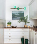 Interior with Dope Gold Long Strip 3 Light Globe Vanity Wall Sconce With Green Glass Shades above white cabinets.