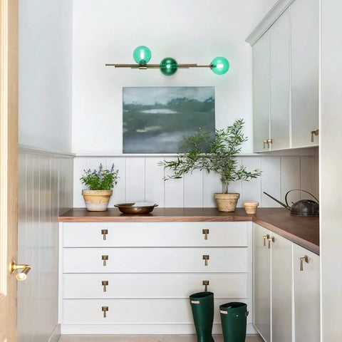 Interior with Dope Gold Long Strip 3 Light Globe Vanity Wall Sconce With Green Glass Shades above white cabinets.