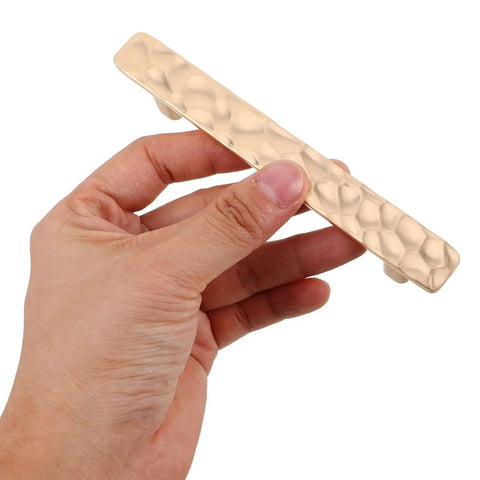 Hand holding a gold zinc alloy cabinet pull handle from Two Pack Zinc Alloy Cabinet Pull Handles With Screws For Kitchen Cabinets Wardrobe And Dresser Doors Modern Metal Drawer Pulls Hardware Accessories Dope Quality Ready To Install Built To Last. Focus on textured surface.