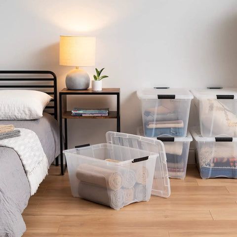 See Through 53 Quart Stackable Plastic Storage Bin with Lid for Home Organization, towels inside, beside bed and lamp.