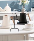 Black metal coffee dripper stand beside white and black kettles on a table, showcasing Grab A Fresh Cup With This Premium Solid Metal Coffee Dripper Stand Perfect Drip Rack Station For Hand Pour Brewers Filter Holders Stable Anti Tip Base Makes Daily Brewing Sessions Easy Whether You Chill At Home Or Travel Light.