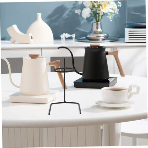 Black metal coffee dripper stand beside white and black kettles on a table, showcasing Grab A Fresh Cup With This Premium Solid Metal Coffee Dripper Stand Perfect Drip Rack Station For Hand Pour Brewers Filter Holders Stable Anti Tip Base Makes Daily Brewing Sessions Easy Whether You Chill At Home Or Travel Light.