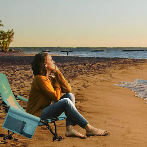 Person sitting on low beach chair for beach tent and camping, ultralight folding recliner with cup holder by the shore.