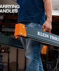 Carpenter carrying Tough As Nails Folding Sawhorses with orange handles in garage setting.