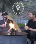 Two friends enjoying ice cream cones near a campfire, surrounded by nature, with a tent in the background.